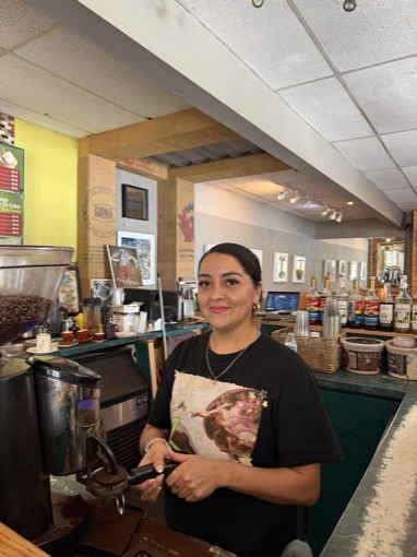 A barista smiling behind the espresso machine next to the coffee grinder