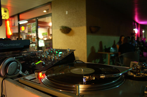 A DJ setup with a turntable and mixer on the patio at night, with the lit-up shop and customers behind it