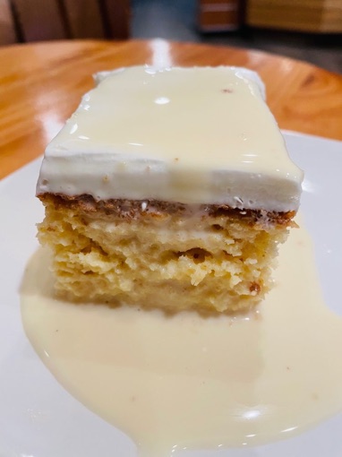 A square of tres leches cake topped with whipped cream, sitting in a pool of sweet milk
